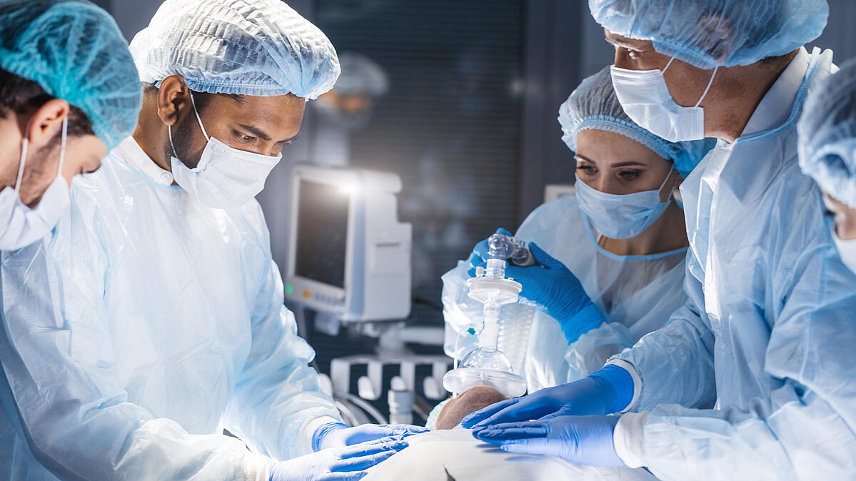 OP-Saal mit Ärzten die sich über einen Patienten beugen, mittig Medizinfilter über der Nase zm Beatmen