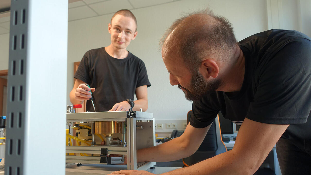 Auszubildener schaut vom Werkstück auf, im Vordergrund weiterer Ingenieur bei der Arbeit