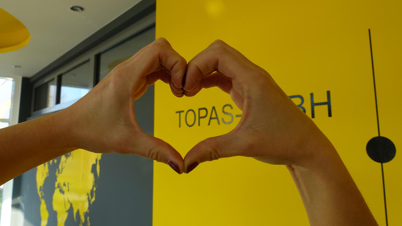 Eingangsbereich Topas GmbH mit Weltkarte und Firmenname, davor Hände die ein Herz vor dem Namen Topas formen