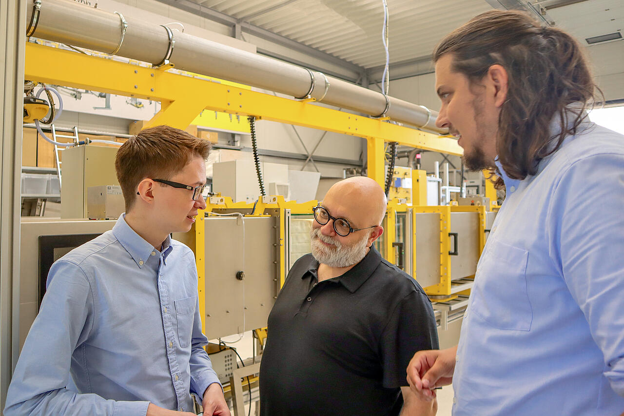 Produktionshalle mit Pr&uuml;fstand, davor drei Personen im Gespr&auml;ch