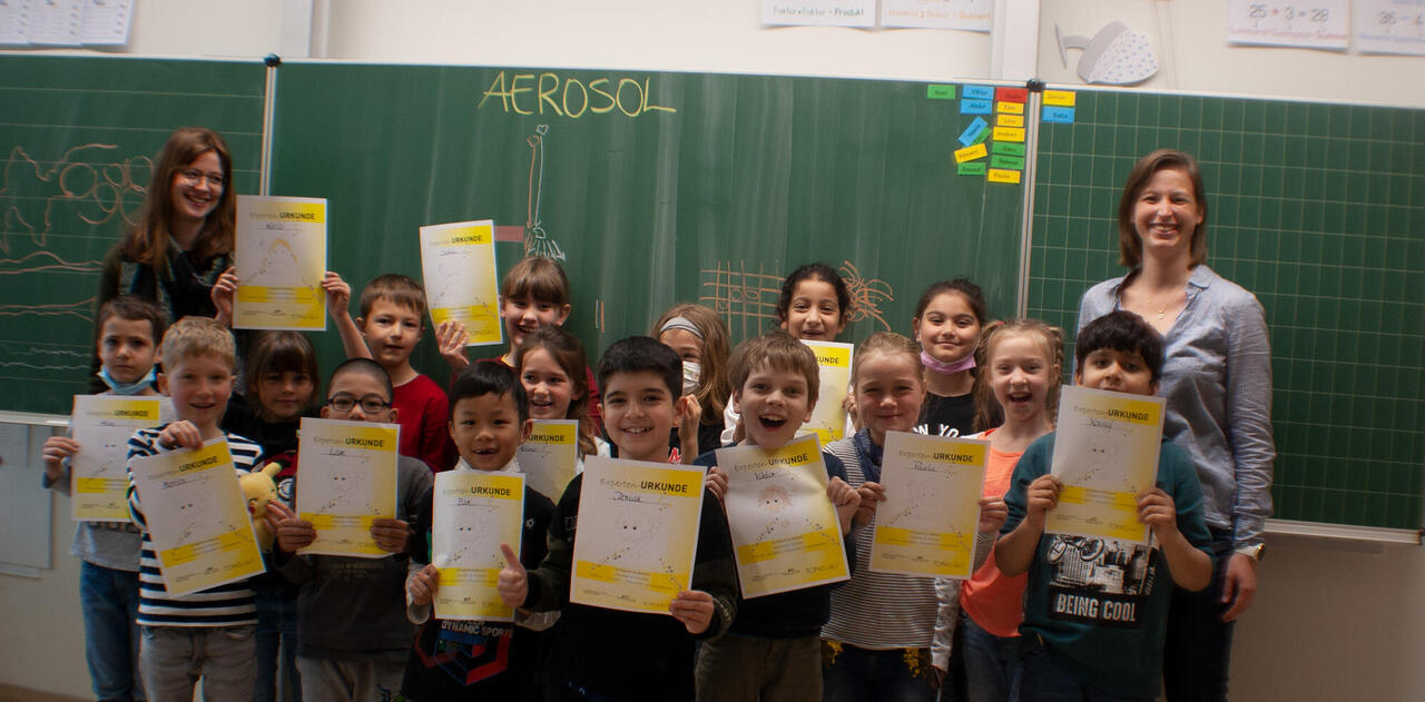 Lachende Schulklasse versammelt vor der Tafel mit Urkunden in der Hand und "Aerosol" an der Tafel geschrieben