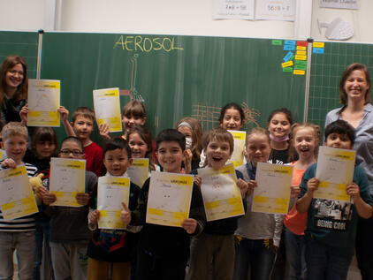 Lachende Schulklasse versammelt vor der Tafel mit Urkunden in der Hand und "Aerosol" an der Tafel geschrieben