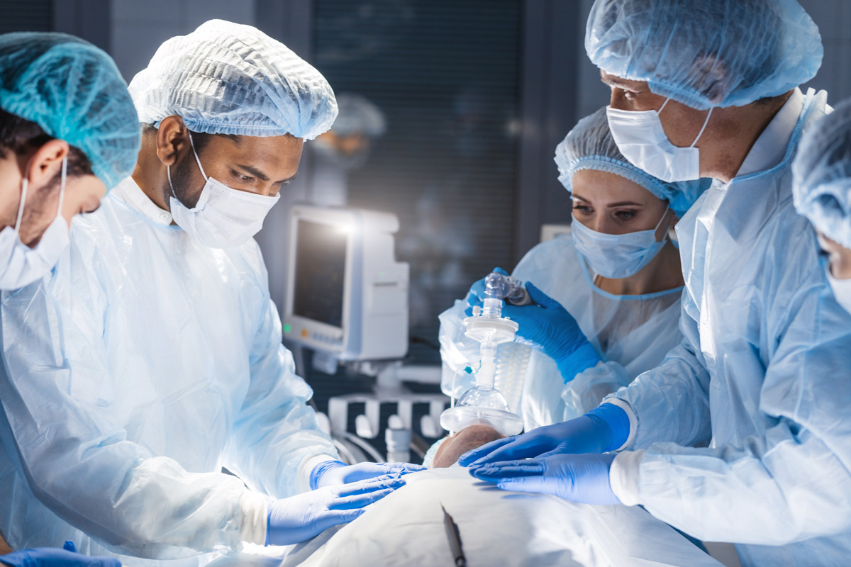 OP-Saal mit Ärzten die sich über einen Patienten beugen, mittig Medizinfilter über der Nase zm Beatmen